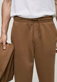 Pantaloni de trening maro, cu talie elastică, închidere cu șnur și buzunare laterale. Materialul este moale și ușor texturat.