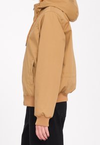 Vue de côté d'une personne portant une veste à capuche beige avec des poignets et un ourlet côtelés, associée à un pantalon noir sur un fond blanc.