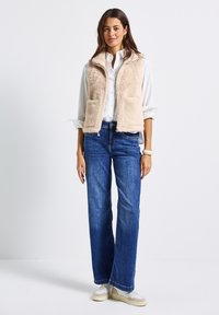 Gilet in pelliccia sintetica beige con due tasche anteriori, indossato su una camicia bianca con bottoni. Abbinato a jeans blu a gamba dritta e sneaker chiare.