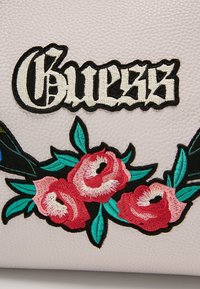 Surface blanche texturée avec le texte brodé en noir et blanc « Guess » au-dessus de trois roses rouges et roses avec des feuilles vertes.