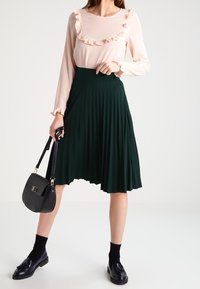 Vaaleanpunainen pitkähihainen paita, jossa on röyhelökaulus, tummanvihreä poimutettu hame, musta käsilaukku, mustat sukat ja kiiltävät mustat loaferit.