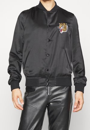 Homme portant une veste bomber en satin noir avec une tête de tigre brodée sur la poitrine et un pantalon en cuir noir avec une fermeture éclair sur le devant.