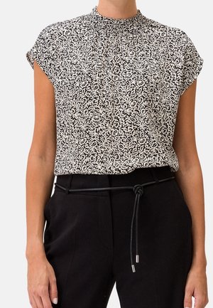 Kvinde iført sorte bukser med tyndt sort bælte og hvid og sort abstrakt mønstret ærmeløs bluse med høj hals.