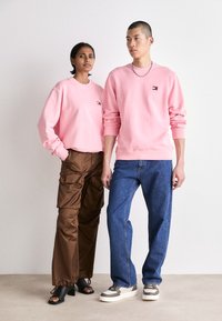 Dos personas que llevan sudaderas rosas con un pequeño logo, combinadas con pantalones cargo marrones y jeans azules, de pie sobre un fondo neutro.