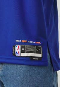 Blå mesh basketballtrøje med åndbart stof, et mærke med "SWINGMAN," NBA-logo og størrelse 44. Teksten inkluderer "ONCE A KNICK, ALWAYS A KNICK."