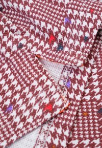 Textiel met een houndstoothpatroon in roestkleur en wit, met kleurrijke stippen. Zichtbare stiksels en rafels aan de randen.