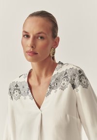 Blouse blanche avec broderie florale noire le long de l'encolure, présentant une ouverture en V et des manches longues. Des boucles d'oreilles dorées texturées ornent le modèle.