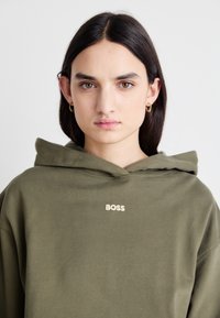Felpa verde oliva con tasca a marsupio, cappuccio con cordini e logo "BOSS" in crema. Stoffa morbida con vestibilità comoda.