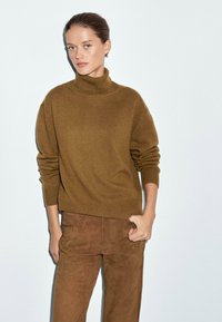 Maglione a collo alto khaki in un morbido tessuto a maglia con maniche lunghe, abbinato a pantaloni in suede marroni. L'outfit ha una vestibilità rilassata e un design minimale.