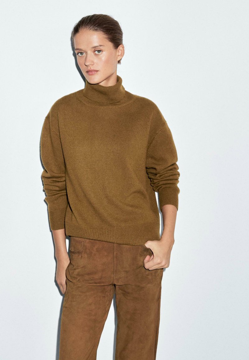 Maglione a collo alto khaki in un morbido tessuto a maglia con maniche lunghe, abbinato a pantaloni in suede marroni. L'outfit ha una vestibilità rilassata e un design minimale.