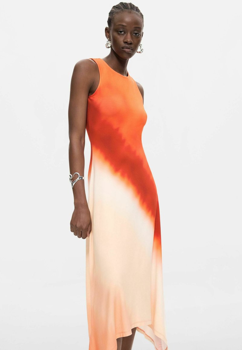 Femme aux cheveux tressés portant une robe sans manches dégradée orange et crème, un bracelet argenté et des boucles d'oreilles créoles, debout devant un fond uni.