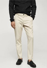 Beige Chinos mit einer schmalen Passform, ausgestattet mit Fronttaschen und einem einzigen Knopfverschluss. Kombiniert mit einem strukturierten schwarzen Pullover und schwarzen Loafers.