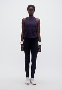 Γυναίκα με σκούρα αθλητικά ρούχα κρατώντας βαράκια χεριών, φορά λευκά βραχιολάκια και λευκά αθλητικά παπούτσια, στέκεται μπροστά σε απλό φόντο.