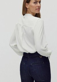 Vit blus med långa ärmar, plisserad bakdel och en liten cirkulär detalj vid halsringningen, kombinerad med mörk denimhögmidjade byxor.