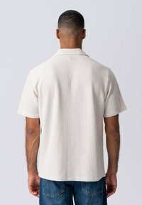 Kortærmet poloshirt i lys beige med tekstureret ribbet stof og klassisk krave, parret med mørkeblå denimjeans.