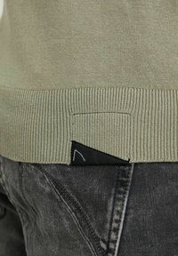 Pull vert olive avec ourlet côtelé, doté d'une étiquette noire et d'un détail de couture subtile sur un jean gris en gros plan.