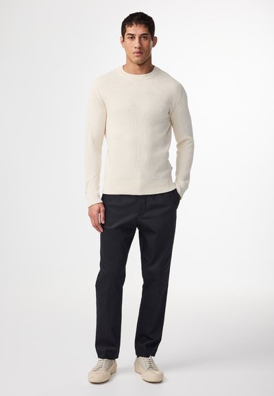 Cremfarbener, gerippter Strickpullover mit rundem Ausschnitt, kombiniert mit dunklen Hosen und hellen Sneakern. Glatte Textur. Einfaches Design.