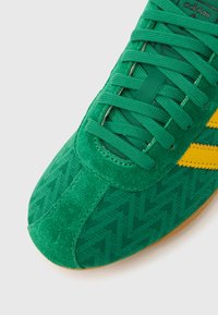 Πράσινο αθλητικό παπούτσι με υφασμάτινη υφή και λεπτομέρειες από σουέτ, διαθέτει επικάλυψη με σχέδιο και δύο κίτρινες ρίγες στο πλάι.