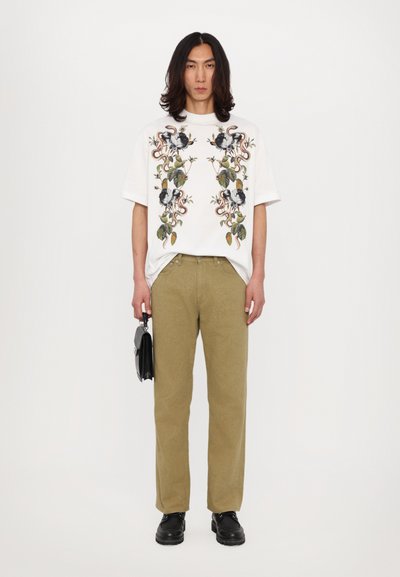 Hvid grafisk t-shirt med blomster- og snakesdesign, parret med lysegrønne bukser og sorte sko, mens der holdes en lille sort taske.