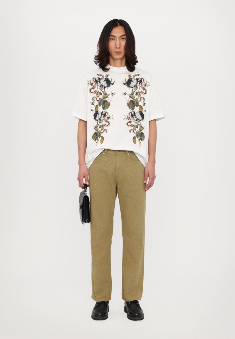 Maglietta grafica bianca con design floreale e di serpente, abbinata a pantaloni verde chiaro e scarpe nere, mentre tiene una piccola borsa nera.