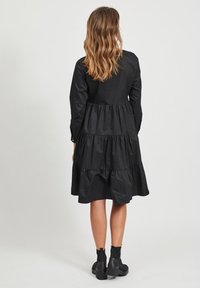 Schwarzes, gestuften Kleid aus leichtem Stoff, mit langen Ärmeln und knielangem Saum. Getragen mit klobigen, schwarzen Stiefeletten.