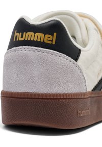 Weißer Sneaker mit grauem Wildlederabsatz, schwarzem Lederakzent und brauner, strukturierter Sohle. "Hummel"-Logo in Gold auf der Rückseite.