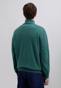 Homme portant un pull col roulé vert et un jean bleu foncé, de dos, sur un fond gris clair uni.