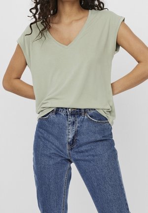 Woman wearing sage green V-neck short-sleeve top tucked into high-waisted blue denim jeans, hands placed on her hips.