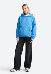 Sudadera azul de gran tamaño con un bolsillo frontal, combinada con jeans oscuros de corte holgado y zapatillas blancas. Textura de tela suave.