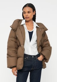 Brun pufferjakke med høj krave, to lynlåslommer og sølvhardware, båret over en hvid stribet skjorte og mørke jeans.