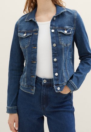 Kvinde iført en åben blå denimjakke over en hvid top og matchende blå højtaljede jeans, med den ene hånd i lommen.
