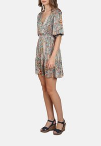 Femme portant une robe courte à motif paisley coloré avec des manches bouffantes et des sandales compensées bleu marine, se tenant contre un fond uni.