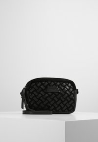 Pieni musta nahkainen rannekassi, jossa on kudottu kuvio ja vetoketjukiinnitys, esillä valkoisella jalustalla yksinkertaista taustaa vasten.