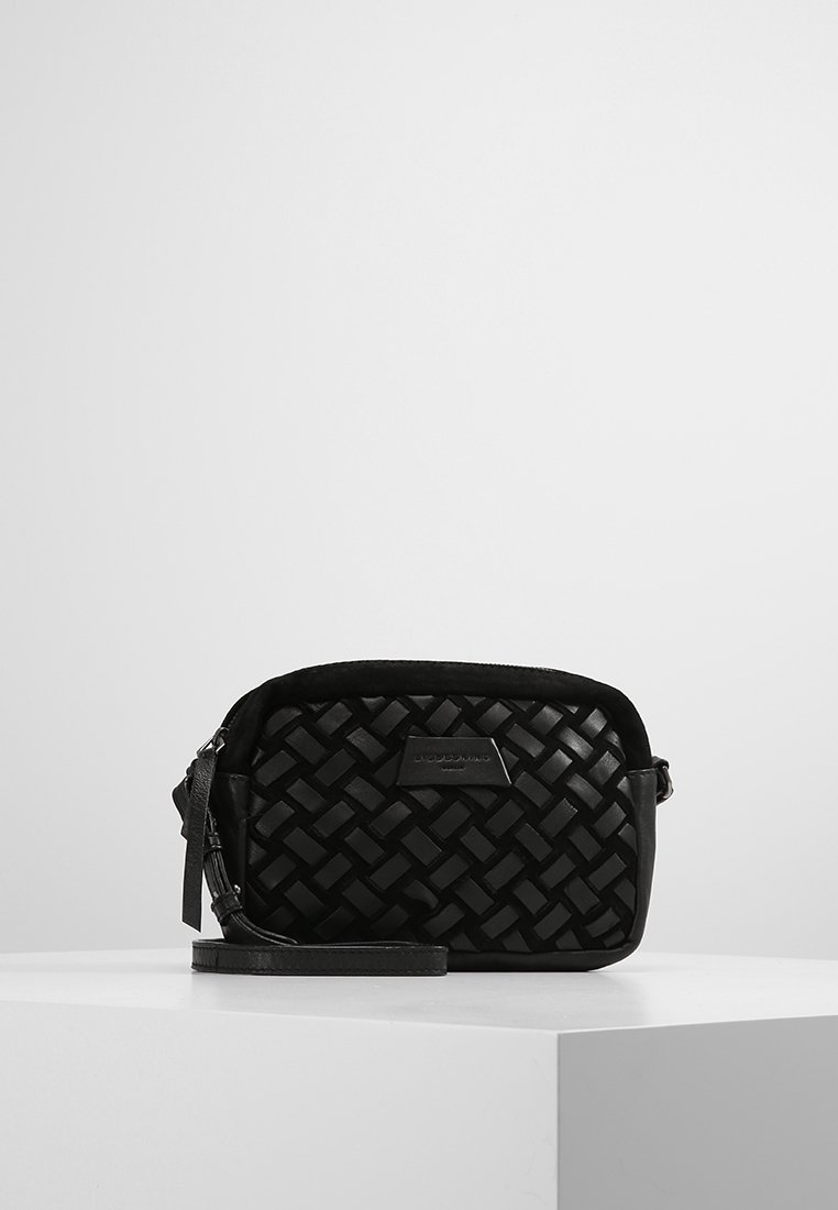 Pieni musta nahkainen rannekassi, jossa on kudottu kuvio ja vetoketjukiinnitys, esillä valkoisella jalustalla yksinkertaista taustaa vasten.