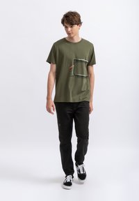 T-shirt en coton vert olive avec une poche et un graphique abstrait. Associé à un pantalon noir avec des accents de couture et des baskets blanches.