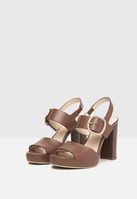 Brown leather platform heels with peep-toe, ankle strap, and square buckle. Chunky block heel with a smooth texture and contrasting beige sole.