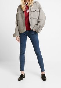 Chaqueta gris de pata de gallo con dos bolsillos en el pecho, sudadera con capucha roja con logo, vaqueros ajustados azules y zapatos negros sin cordones. Modelo posando de forma casual.