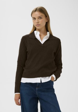 Pull en tricot côtelé marron avec un col en V et un design col, superposé sur une chemise blanche, associé à un pantalon en denim bleu.