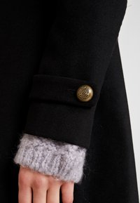 Ręka w szarym dzianym rękawie pod czarnym rękawem płaszcza z ozdobnym mosiężnym guzikiem na mankiecie.