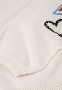 Cremfarbener Pullover mit gerippten Bündchen und Saum, der gestickte Muster eines Herzens und geometrischer Formen in blauen, braunen und schwarzen Fäden zeigt.