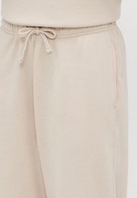Beige joggingbroek met een elastische tailleband en een gestrikt koord, die het bovenbeen en de heupen laat zien.