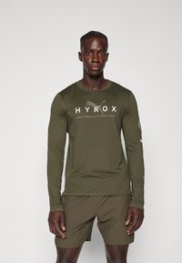 Olijfgroene sportshirt met lange mouwen en witte "HYROX" logo en tekst. Zachte stof, strakke pasvorm, met logo-accenten op de mouwen.