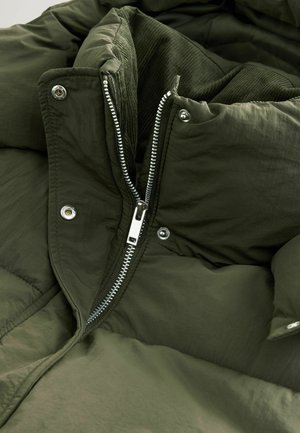 Olivengrøn pufferjakke med en fremtrædende sølvlynlås, trykknapper og et struktureret indre foer med et corduroy-design.