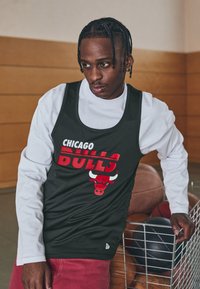 Fekete sporttanktopp, rajta nagy piros betűkkel "CHICAGO BULLS" felirattal és egy piros bika logóval. Fehér hosszú ujjú póló fölött viselve, textúrált felülettel.