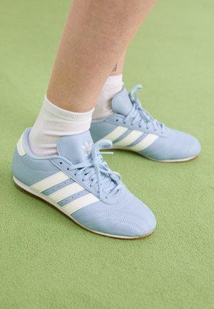 Person iført lyseblå Adidas-sneakers med hvide striber og hvide sokker, der står på en grøn tekstureret overflade.