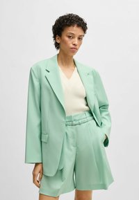 Set blazer verde menta chiaro e pantaloni corti su misura, abbinato a un maglione crema con scollatura a V. Tessuto liscio, vestibilità oversize e design strutturato.