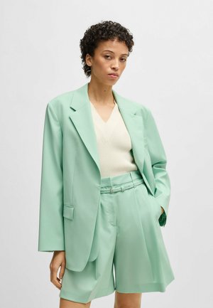 Set blazer verde menta chiaro e pantaloni corti su misura, abbinato a un maglione crema con scollatura a V. Tessuto liscio, vestibilità oversize e design strutturato.