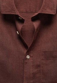 Braunes Leinenhemd mit spitzem Kragen, zwei sichtbaren Knöpfen und einer Brusttasche auf der rechten Seite.