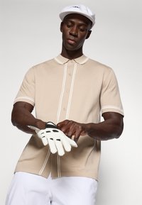 Kreminė polo marškinėliai su balta apdaila, trumpomis rankovėmis, mygtukų uždarymu. Modelis dėvi baltą kepurę ir prisitaiko baltą golfą pirštinę su juodais akcentais.