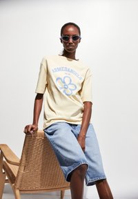 Gelbes Baumwoll-T-Shirt mit blauer Blumenillustration, kombiniert mit blauen Jeansshorts. Das Modell trägt eine helle Sonnenbrille und sitzt auf einem Korbstuhl.
